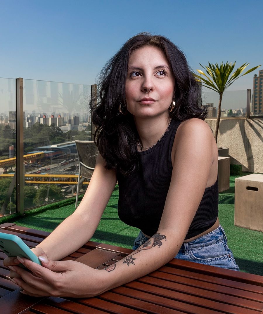 Colaboradora sentada à mesa com o celular, no escritório da Tenda Construtora. Uso institucional.