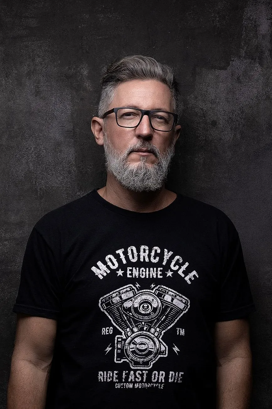 Retrato de Alessandro Couto, fotógrafo, de óculos e barba grisalha, em fundo cinza escuro